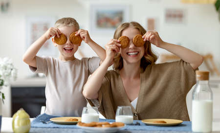 Happy woman and boy covering eyes with oatmeal biscuits while sitting at table and having breakfast in morning in kitchenの写真素材