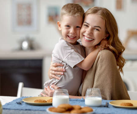 Optimistic woman embracing cheerful boy and looking at camera while sitting at table with cookies and milk and having breakfast in morning at homeの写真素材
