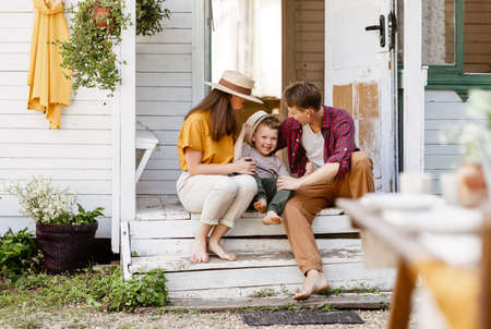 Full body barefoot parents and son smiling while sitting on weathered porch step and relaxing outside house on summer day in countrysideの写真素材