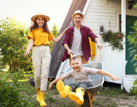 Happy mother and son with outstretched arms smiling and riding wheelbarrow near father outside cottage in summerの写真素材