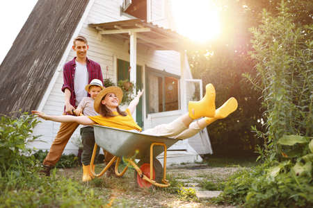 Happy mother and son with outstretched arms smiling and riding wheelbarrow near father outside cottage in summerの写真素材