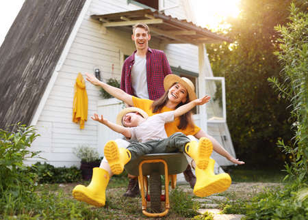 Happy mother and son with outstretched arms smiling and riding wheelbarrow near father outside cottage in summerの写真素材