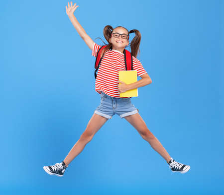 Full body of happy energetic primary schoolgirl wearing casual clothes holding backpackand copybook in hands and jumping high against blue backgroundの写真素材