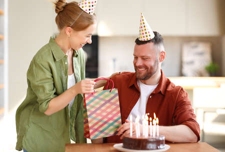 Young positive wife making surprise for husband, excited man holding gift package and being so happy while sitting in kitchen at table with cake. Family couple in party hats celebrating birthday at homeの写真素材