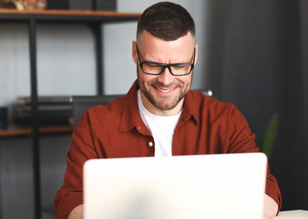 Successful male freelancer in eyeglasses working on laptop computer remotely, man in casual wear...