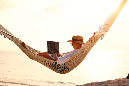 Young woman, successful female freelancer using laptop while lying in hammock on the tropical beach at sunset, working remotely during summer vacation. Distance work conceptの写真素材