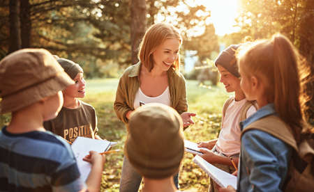 Young positive woman teacher interacting with group of school kids boys and girls during ecology lesson outdoors in forest, kids with notebooks learning about nature and wildlife on sunny autumn dayの写真素材