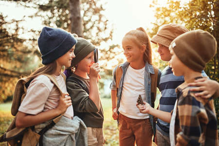 Group of curious happy school kids in casual clothes with backpacks exploring nature and forest together on sunny autumn day, girl holding magnifying glass and looking at classmates with smileの写真素材