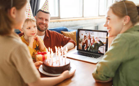 Positive senior couple grandparents wearing party hats congratulating little girl online via video call on laptop, waving at camera and blowing whistles, child celebrating birthday with family at homeの写真素材