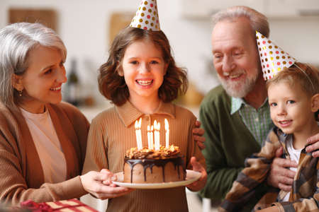 Little girl celebrating birthday while her senior grandparents smiling, child preparing to blow candles on cake and make wish on Birthday, elderly grandma and grandpa congratulating granddaughterの写真素材