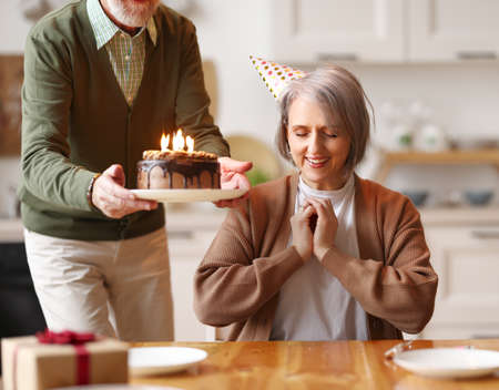 Happy elderly woman in party hat closing her eyes, she makes a wish, while her husband holds a chocolate cake with candles celebrating her Birthday at home, sitting together in kitchenの写真素材