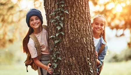 Two happy smiling school girls with backpacks hugging big tree in wild forest and looking at camera during camping activity in nature on warm sunny day. Children exploring environment outdoorの写真素材