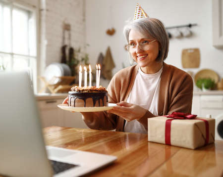 Beautiful happy senior woman with chocolate cake with candles in hands, celebrating birthday online with family, looking at web cam on laptop, receiving congratulations during self isolation at homeの写真素材