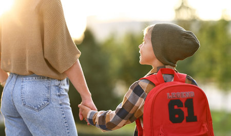 Rear view of mother and little son schoolboy with backpack holding hands, going to school together...
