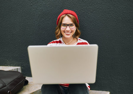 Photo of smiling student girl in glasses wearing earphones sitting outside with open laptop looking...