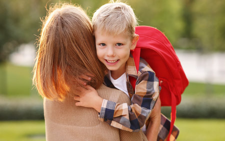 Ready to study. Cute happy little schoolboy with backpack hugging embracing his mother before going...