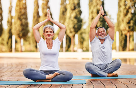 Calm elderly retired family couple practicing yoga outside happy husband and wife sitting on mats barefoot with eyes closed in lotus position with folded hands raised up. Meditation and mental healthの写真素材