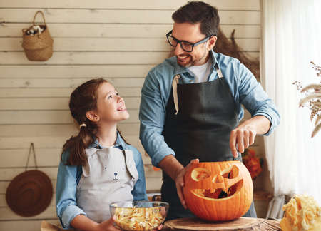 Smiling father showing his little daughter how to carve pumpkin with spooky face, dad with child creating this iconic symbol of Halloween holiday jack-o-lantern while standing behind table in kitchenの写真素材