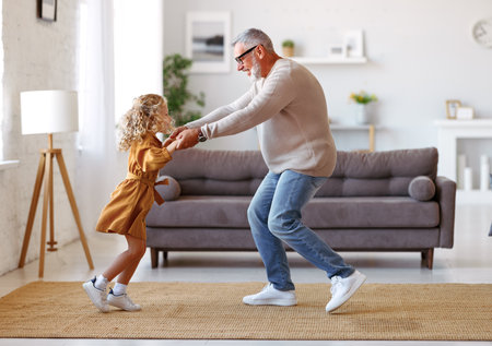 Active senior man grandfather enjoying dance with cute little granddaughter in stylish living room at home, small adorable girl in dress dancing with grandpa while spending time together on weekendの写真素材