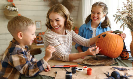 Positive young family mother with children son and daughter smiling while creating jack-o-lantern during Halloween celebration, happy mom and kids drawing on pumpkin in kitchen at homeの写真素材