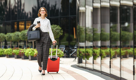 Ready for business trip. Full length of young happy stylish business woman in formal outfit with suitcase luggage walking along city street and smiling, holding take away coffee and handbag in handの写真素材