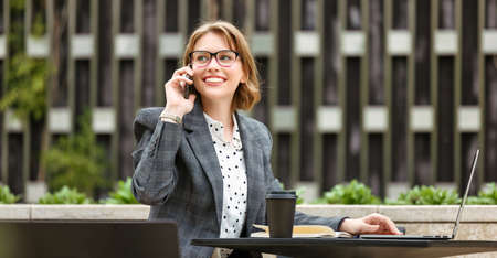Smiling attractive businesswoman in formal outfit talking on mobile phone while waiting for partner or client at meeting in outdoors cafe, sitting at table with opened laptop and making callの写真素材