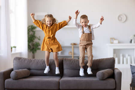 Full length of overjoyed little children brother and sister jumping on sofa, laughing and having fun while playing together at home, kids enjoying carefree leisure time at homeの写真素材