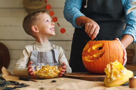 Cute happy little boy helping his father to carve Halloween pumpkin while standing in kitchen at home and preparing for autumn holiday, family of two son and dad making Jack-o-Lantern togetherの写真素材