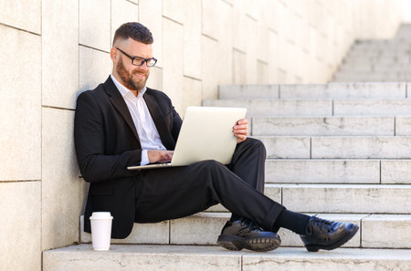 Young happy businessman freelancer in suit sitting on stairs outside with laptop and coffee to goの写真素材