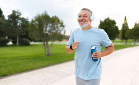 Happy mature man in headphones holding water bottle while jogging outside in park in early morning, senior sportsman running with beaming smile on his face enjoying active healthy lifestyleの写真素材