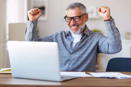 Excited senior man celebrating success, getting good exam results during distant education, raising hands with clenched fists and looking at laptop with happy face expression while sitting at deskの写真素材