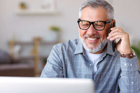 Handsome cheerful senior man wearing glasses talking on mobile phone and looking at laptop, working remotely at home, elderly businessman enjoying pleasant conversation with client at his workplaceの写真素材