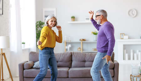 Energetic senior family couple dancing together in living room at home, moving to music and smiling, positive retired people wife and husband enjoying active life on retirementの写真素材