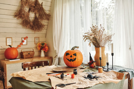 Happy Halloween. Carved orange pumpkin with drawn spooky face standing on wooden table in cozy rustic kitchen, classic Jack-o-lantern in house interior during holiday preparationの写真素材