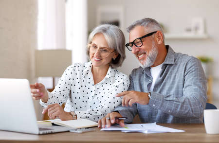 Senior family couple studying online together, retired man and woman looking at laptop and discussing something while sitting at desk at home, enjoying distant education on retirementの写真素材