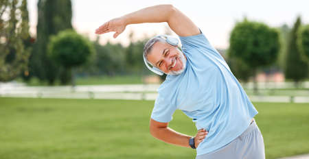 Staying active after retirement. Happy joyful mature retired sportsman wearing headphones and sportswear doing side stretching exercises with arm over his head, exercising outside in city parkの写真素材