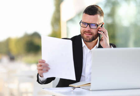 Focused serious businessman in formal suit talking by mobile phone with client and looking at document paper, reading contract while sitting at outdoors cafe and working onlineの写真素材