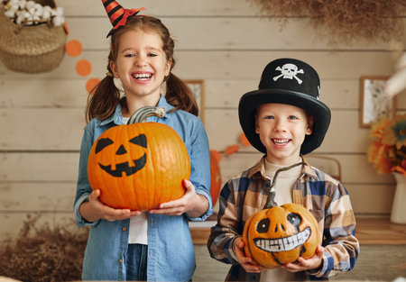 Cute happy children sister and brother holding carved orange pumpkins with drawn funny faces while preparing for family Halloween party at home, kids showing Jack-o-lanterns at cameraの写真素材
