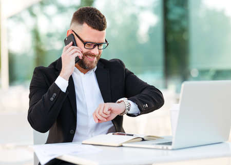 Smiling businessman in formal suit making call by mobile phone and checking time on hand watch, waiting for business partner at meeting in outdoor cafe, entrepreneur working remotely on laptop outsideの写真素材