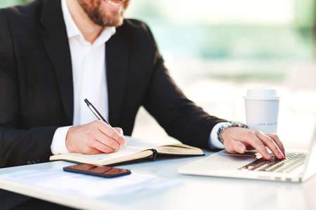 Cropped shot of businessman in suit working remotely on laptop computer at outdoors cafe, watching webinar and taking notes during coffee break, entrepreneur freelancer sitting outsideの写真素材