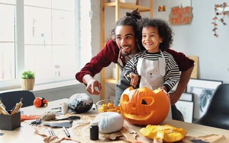 Smiling african american parent father removing pulp from ripe pumpkin while carving jack o lantern with little son for Halloween celebration at home in kitchen and looking with smileの写真素材
