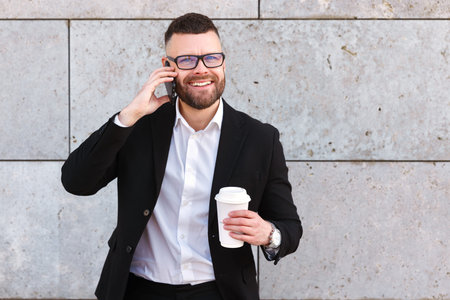 Young positive confident businessman in formal wear speaking on mobile phone with take away coffee in hand, standing against concrete wall of city building, office worker with smartphone outsideの写真素材