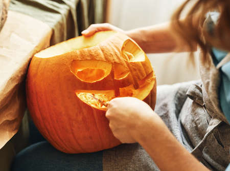 Cropped shot of female carving large orange pumpkin for Halloween party while sitting at wooden table at home, making scary face on jack-o-lantern with knife to set the mood for trick-or-treatersの写真素材