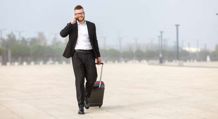Full length of young confident businessman in formal black suit pulling suitcase luggage and talking by phone after landing in airport, calling for taxi. Business trip conceptの写真素材