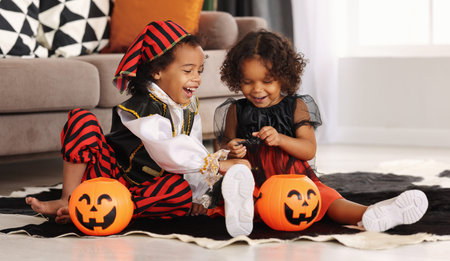 Two cute african kids brother and sister in Halloween costumes playing with artificial spider toy while sitting on floor with orange pumpkin lantern during All Saints Day Party at homeの写真素材