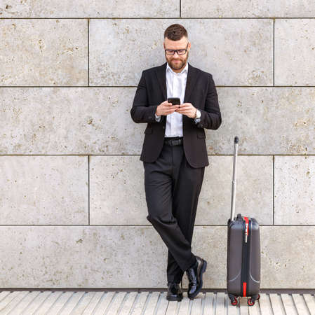 Full length of young confident businessman in formal outfit with suitcase luggage using mobile phone outdoors, waiting for taxi while standing against concrete wall outsideの写真素材