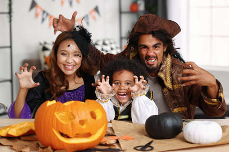 Joyful happy african american family parents and little boy son in Halloween costumes making scary gesture at camera while making jack o lanternの写真素材