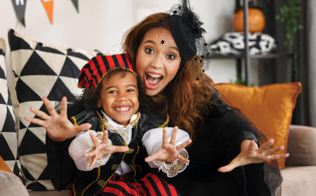 Joyful african american family mother and little boy son in Halloween costumes making scary gesture and looking at camera while sitting on sofa in decorated living room, celebrating all hallows dayの写真素材