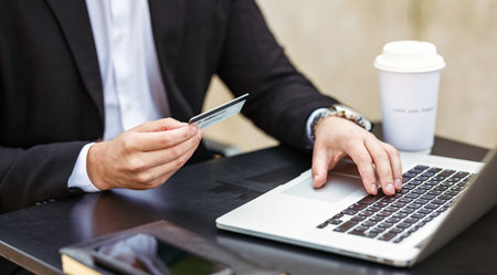 Cropped shot of business professional man holding in hand credit bank card and paying online while sitting at cafe table with laptop, using internet banking during freelance work outsideの写真素材