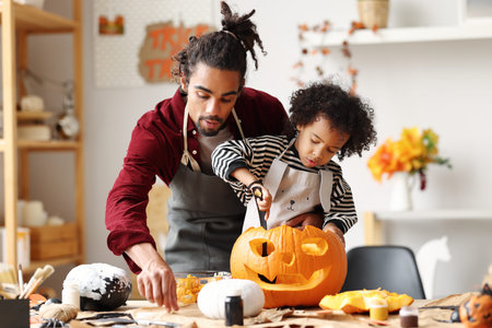 Cute happy little african american boy carving Halloween pumpkin with father at home, mixed-race family son and dad making Jack-o-Lantern together while preparing decorations for Saints Day partyの写真素材